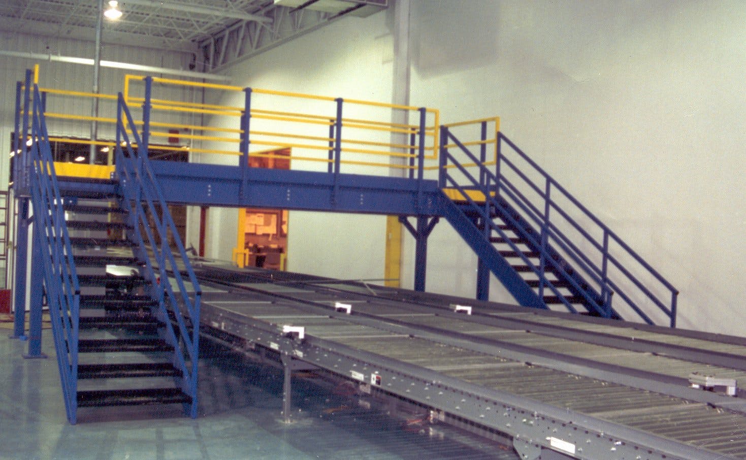 Blue crossover catwalk with OSHA yellow safety handrails above grey shipping conveyor belt