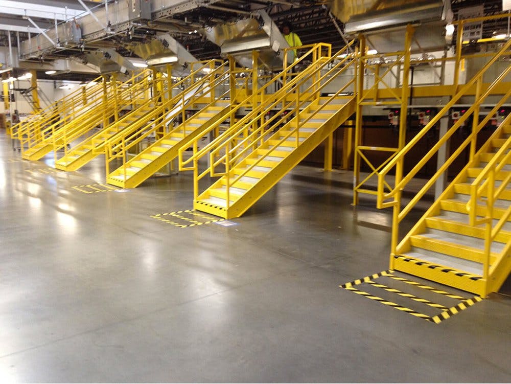 OSHA yellow one stringer stainless steel stairs with safety handrails in manufacturing facility with man in yellow vest at control unit at top of stairs